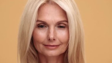 Yüzün yakın plan görüntüsü. Orta yaşlı, gülümseyen bir kadın. Bej stüdyoda kameraya bakıyor.