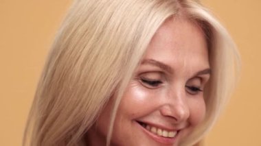 Gülümseyen orta yaşlı kadının mükemmel tenini bej stüdyodaki kameraya gösteren yakın plan bir görüntüsü.