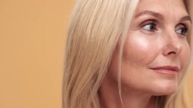 Pozitif orta yaşlı kadının yan tarafa bakıp bej rengi stüdyoda mükemmel tenini gösterişi.