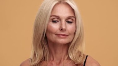 Düşünceli orta yaşlı bir kadın yan tarafa bakıyor ve bej rengi stüdyoda sessizlik jesti yapıyor.