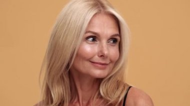 Gülümseyen orta yaşlı bir kadın yan tarafa bakıyor ve bej rengi stüdyoda sessizlik hareketi yapıyor.