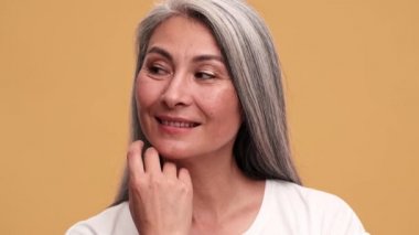 Olumlu Asyalı olgun bir kadın saçını düzeltiyor ve bej renkli stüdyodaki kameraya bakıyor.