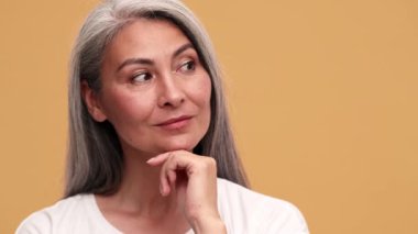 Gülen Asyalı olgun bir kadın bej stüdyoda elini yüzünün yakınında tutuyor.