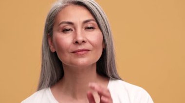 Gülümseyen Asyalı olgun bir kadın kameraya aynaymış gibi bakıyor ve bej rengi stüdyodaki görüntüsünü düzeltiyor.