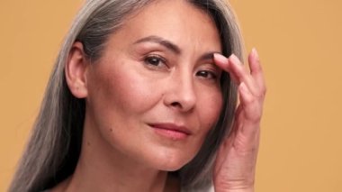 Olumlu Asyalı olgun bir kadının kameraya ayna gibi baktığı ve bej rengi stüdyodaki görüntüsünü düzelttiği yakın plan bir görüntü.