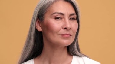 Kendine güvenen Asyalı olgun bir kadının, bej rengi stüdyodaki ayna gibi, kamera karşısında saç stilini düzelttiği yakın plan bir görüntü.