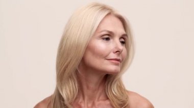 Kendine güvenen olgun, güzel bir kadının yakın çekim görüntüsü. Yanlara bakıyor ve açık bej stüdyodaki kameraya sessiz bir jest gösteriyor.