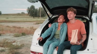 Gülümseyen çok uluslu bir çift doğada bir arabanın bagajında otururken yemek yiyor ve konuşuyor.