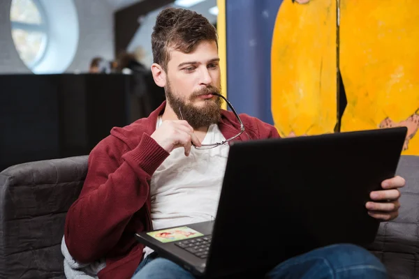 Młody człowiek z broda za pomocą laptopa i trzyma okulary — Zdjęcie stockowe