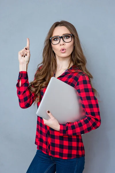 Menina bonita em óculos apontando para cima e segurando laptop — Fotografia de Stock