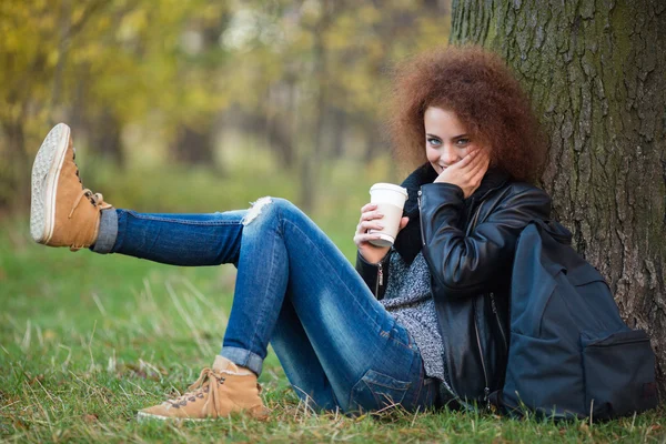 Kobieta siedzi pod drzewem i picia kawy w parku jesień — Zdjęcie stockowe