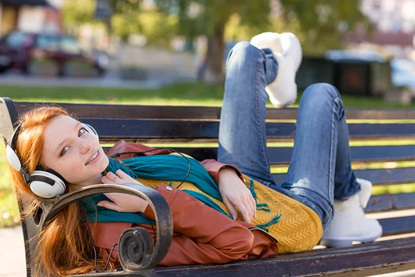 Радостная улыбающаяся девушка отдыхает на скамейке в парке с помощью наушников — стоковое фото