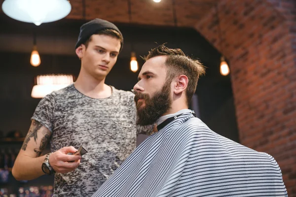 Концентрований перукар робить зачіску бородатому красивому чоловікові в перукарні — стокове фото