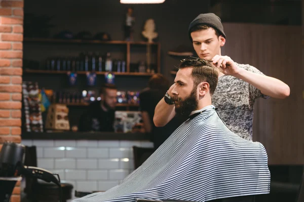 Молодий перукар робить зачіску бородатого чоловіка в перукарні — стокове фото