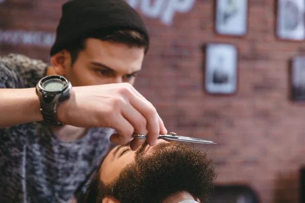 Jovem barbeiro consagrado que prepara a barba do homem com tesoura — Fotografia de Stock