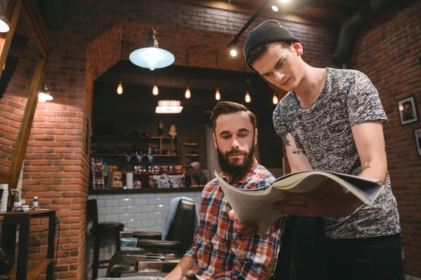 Cabeleireiro jovem e cliente barbudo olhando através da revista — Fotografia de Stock