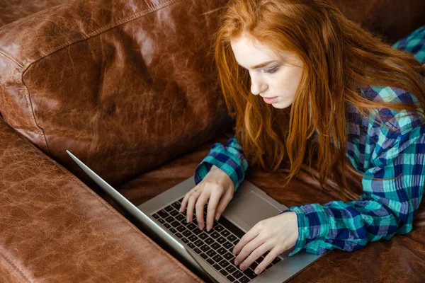 Femme rousse concentrée travaillant avec un ordinateur portable et couchée sur le canapé — Photo