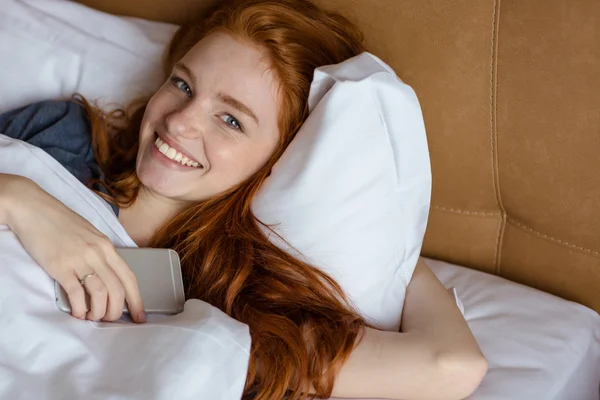Rudy kobietę leżącą w łóżku z smartphone — Zdjęcie stockowe