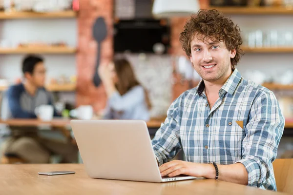 Vrolijke aantrekkelijke jonge krullend man met laptop in café — Stockfoto