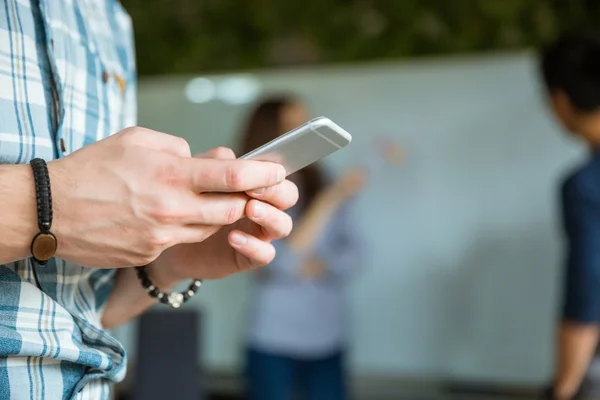 Hände eines jungen Mannes, der sein Smartphone benutzt, während seine Kollegen arbeiten — Stockfoto