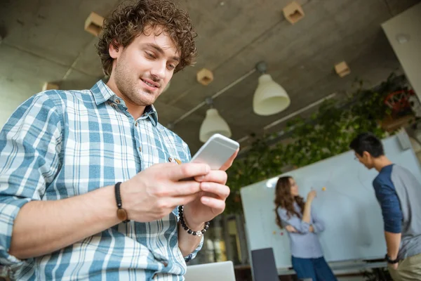 Sorridente uomo riccio utilizzando il cellulare mentre i suoi amici di studio — Foto Stock