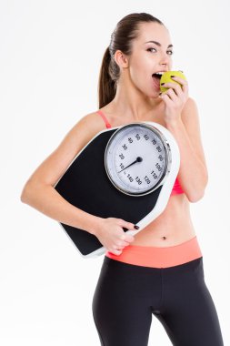 Pretty content sportswoman eating apple and holding weighing scale