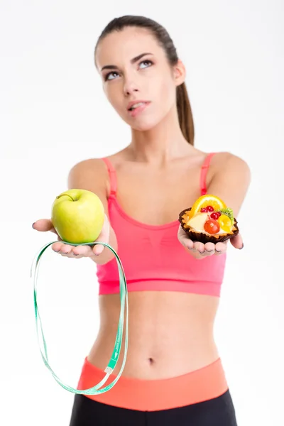 Thoughtful unsure fitness girl holding apple, measuring tape and cake — 스톡 사진