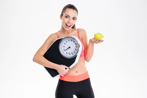 Excited positive fitness girl holding weighing scale and apple — 스톡 사진