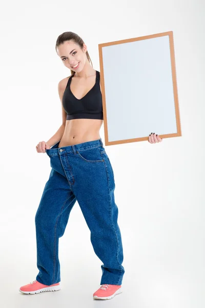 Cheerful young fitness woman in big jeans holding blank board — Stock Photo, Image