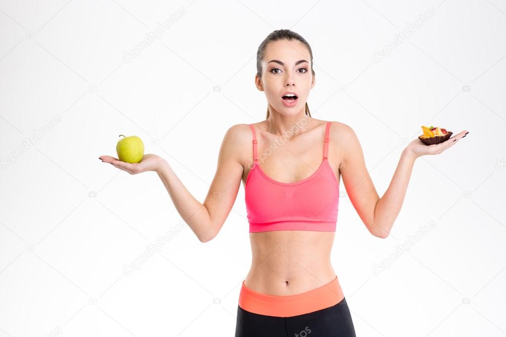 Confused excited young sportswoman choosing between fruits and sweets