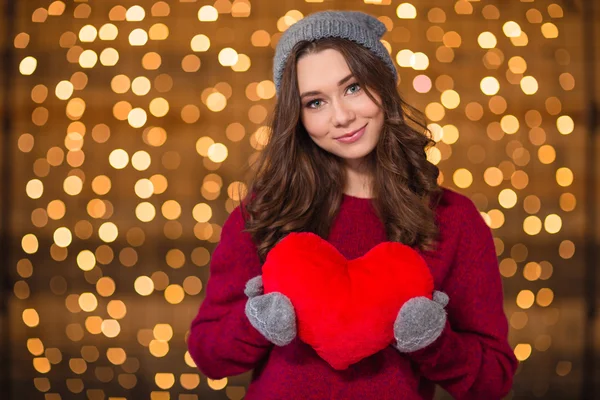 Hermosa mujer feliz sosteniendo el corazón rojo sobre las luces de vacaciones fondo — Foto de Stock