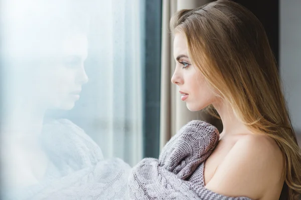 Beautiful woman in grey knitted coverlet looking out of window — Stock Photo, Image