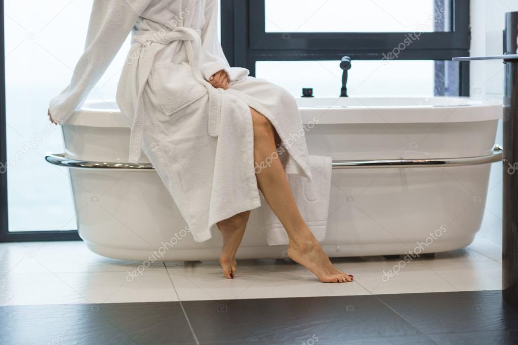 Attractive young barefooted woman in white bathrobe sitting on bathtub 