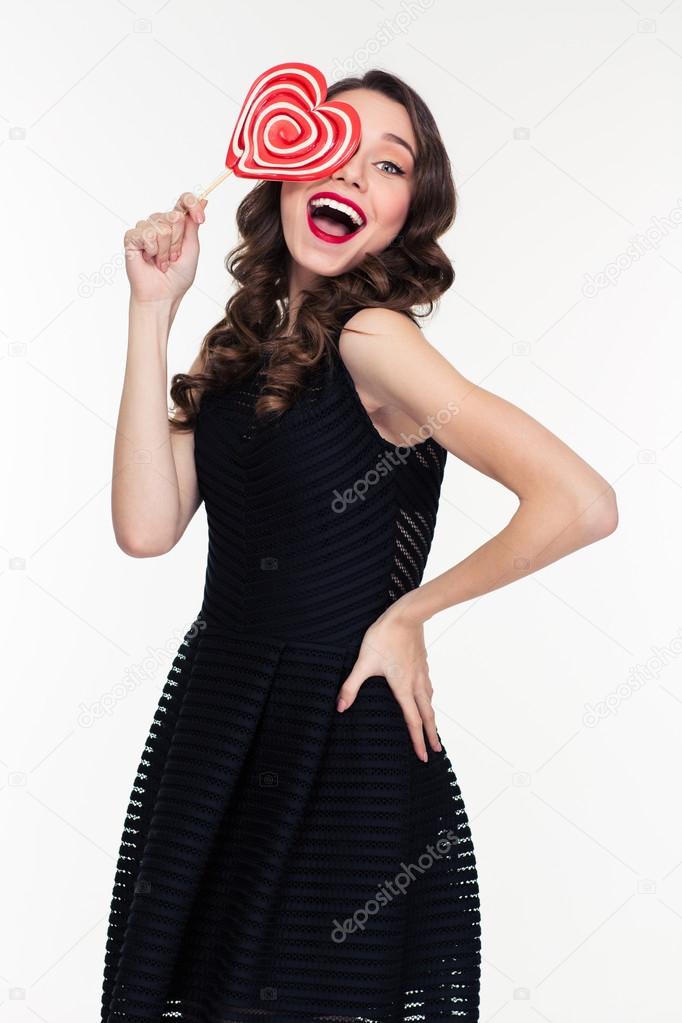 Cheerful charming woman covered her eye with heart shaped lollipop