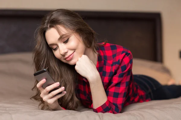 Boldog asszony feküdt az ágyon, és használ smartphone — Stock Fotó