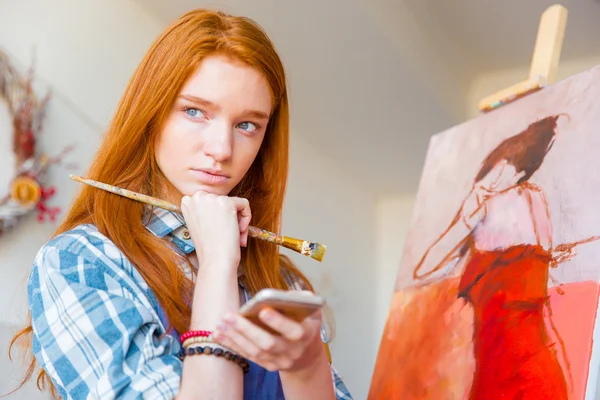 Задумчивая привлекательная художница думает и держит мобильный телефон в мастерской — стоковое фото