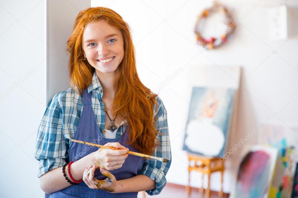 Cheerful beautiful woman painter with long red hair holding brush 