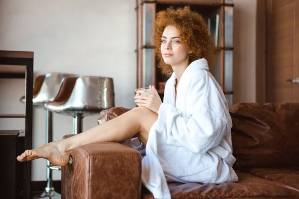 Attractive thoughtful woman in white bathrobe sitting on brown sofa — Stockfoto