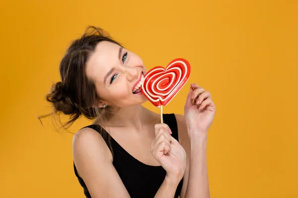 Bonito divertido jovem mulher mordendo doce coração em forma de pirulito — Fotografia de Stock
