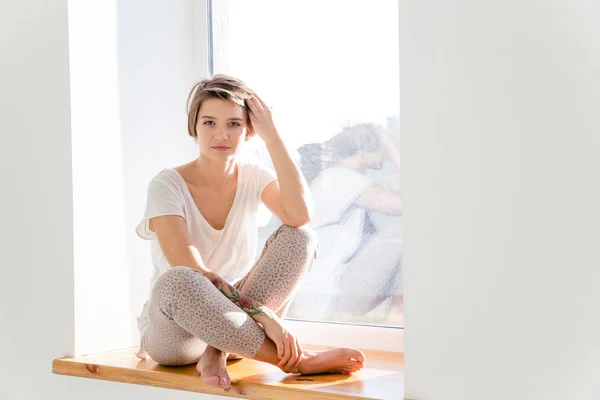 Leuke mooie jonge vrouw in pyjama zittend op de vensterbank — Stockfoto