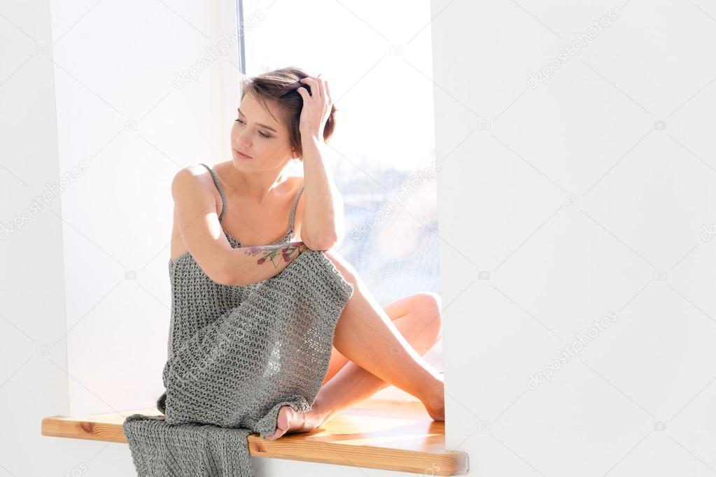 Thoughtful beautiful young woman sitting on window sill and dreaming 