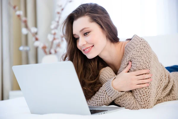 Sonriendo bonita mujer joven acostada en la cama con el ordenador portátil —  Fotos de Stock