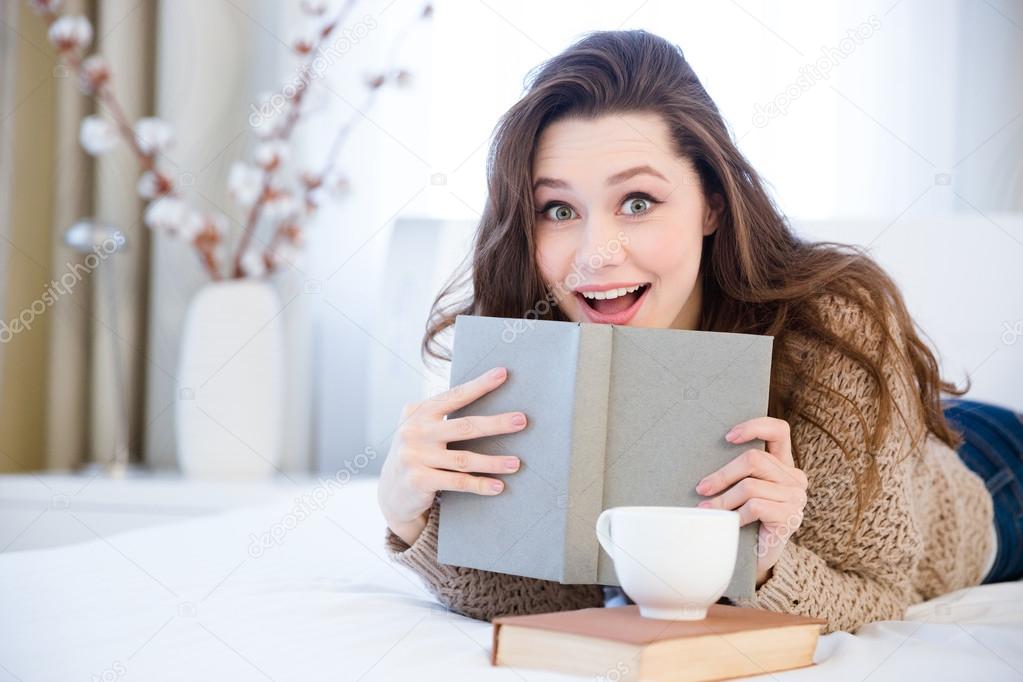 Amazed woman lying on bed and reading book in bedroom
