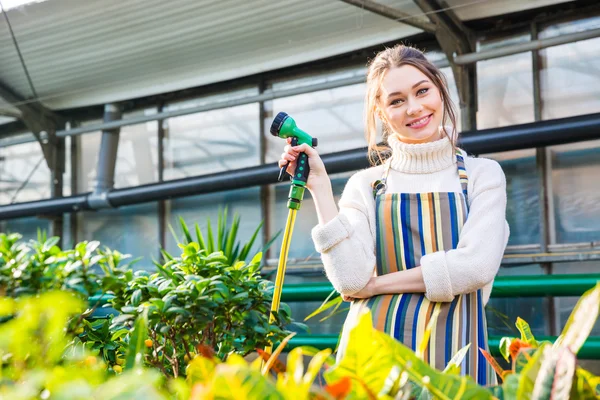 Улыбающаяся женщина, стоящая в оранжерее и держащая садовый шланг — стоковое фото