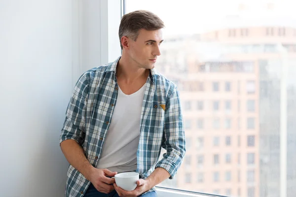 Hombre guapo sentado en el alféizar de la ventana y beber café —  Fotos de Stock