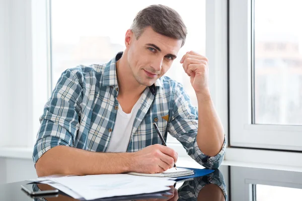 Bonito homem sentado à mesa e escrevendo — Fotografia de Stock