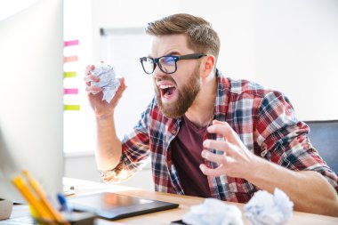 Angry crazy designer yelling and crumpling paper on his workplace clipart