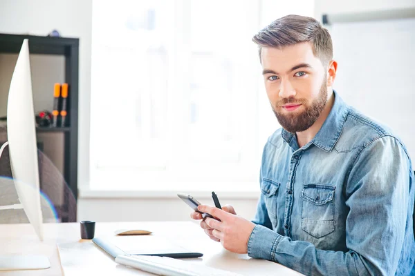 Handsome man sitting on workplace and using mobile phone — Stock Photo, Image