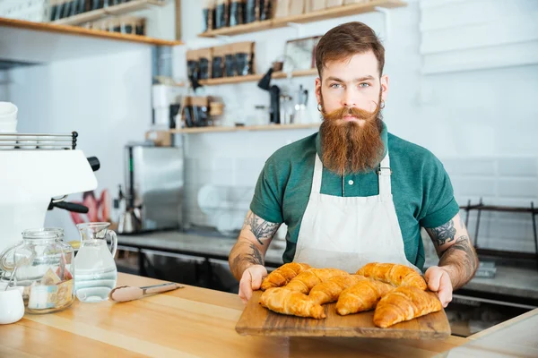 Bearded barista standing and holding wooden board with croissants — Stock Photo, Image