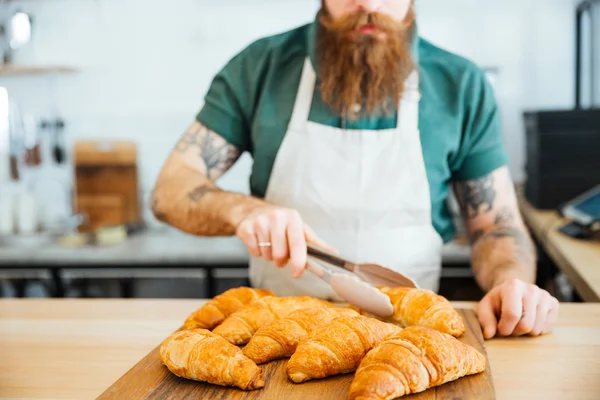 Joven camarero con barba tomando croissant con pinzas —  Fotos de Stock